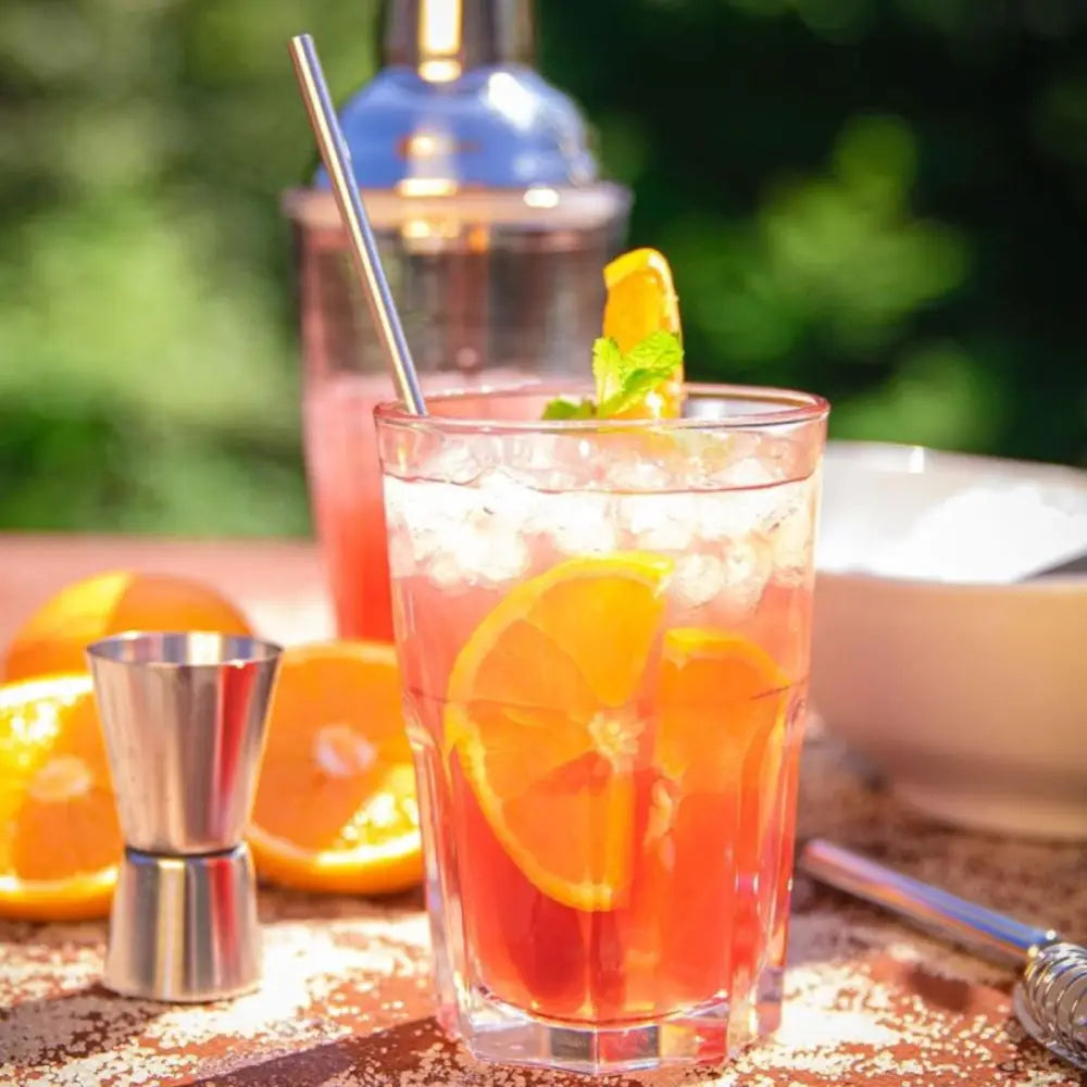 Cocktail with orange slices and ice in a glass, surrounded by oranges and bar tools on a wooden surface.