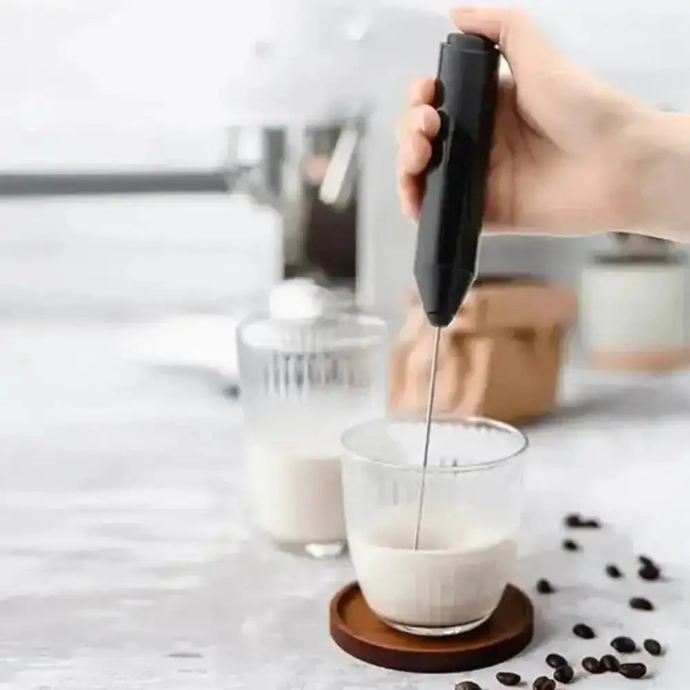 Hand using a milk frother to create foam in a glass of milk with coffee beans and a coffee machine in the background.