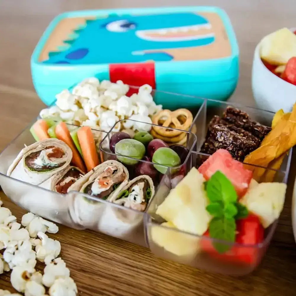 Children's snack box with various food items including popcorn, carrots, grapes, and fruit salad on a wooden surface.