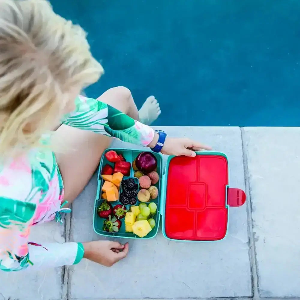 Child sitting by a pool with a red and green lunch box containing fruits.