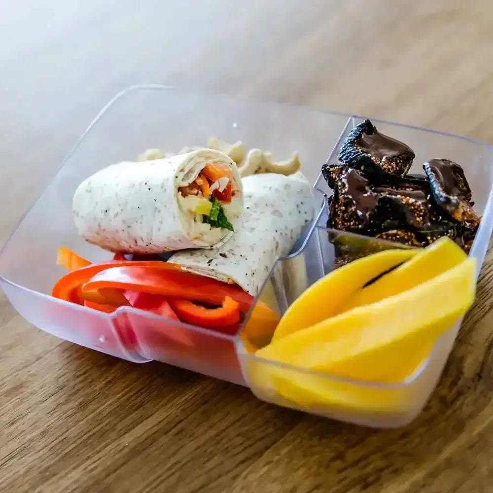 Three-compartment lunchbox with wrap, sliced peppers, chocolate snacks, and mango on a wood table.