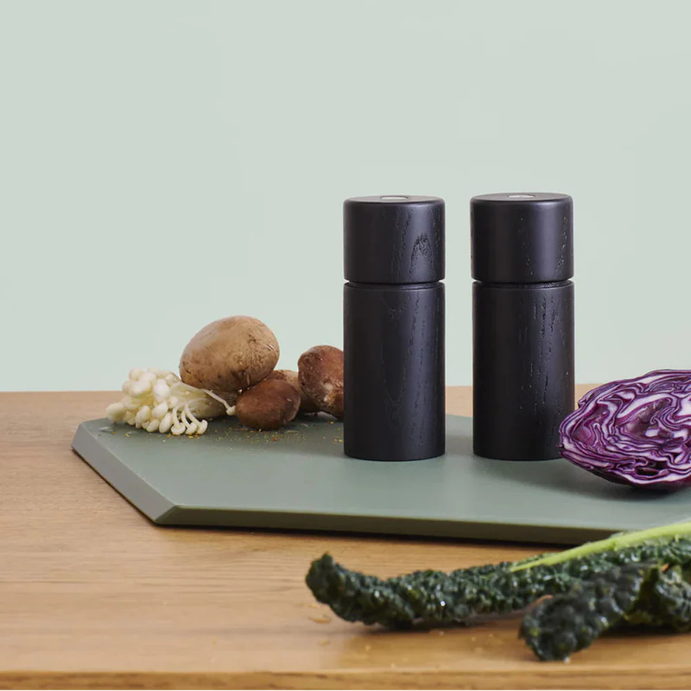 Two black salt and pepper shakers on a green cutting board with vegetables on a wooden surface.