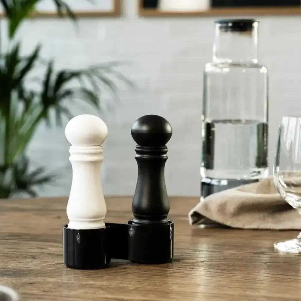 Black and white pepper grinders on a wooden table with a glass of water and napkin in the background.