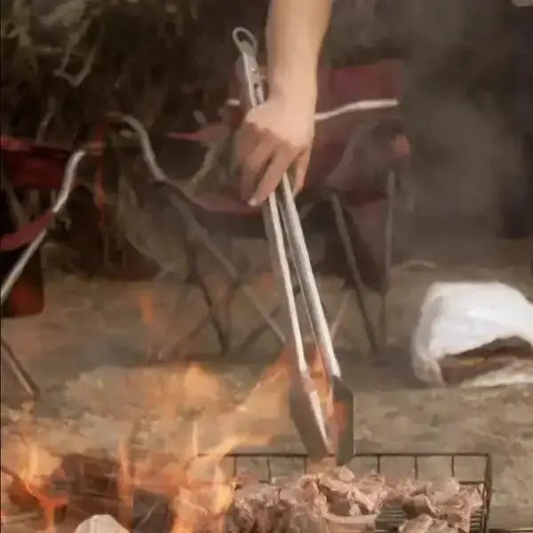 Person using long metal tongs to grill meat over open flame outdoors, Cookin Stores kitchenware