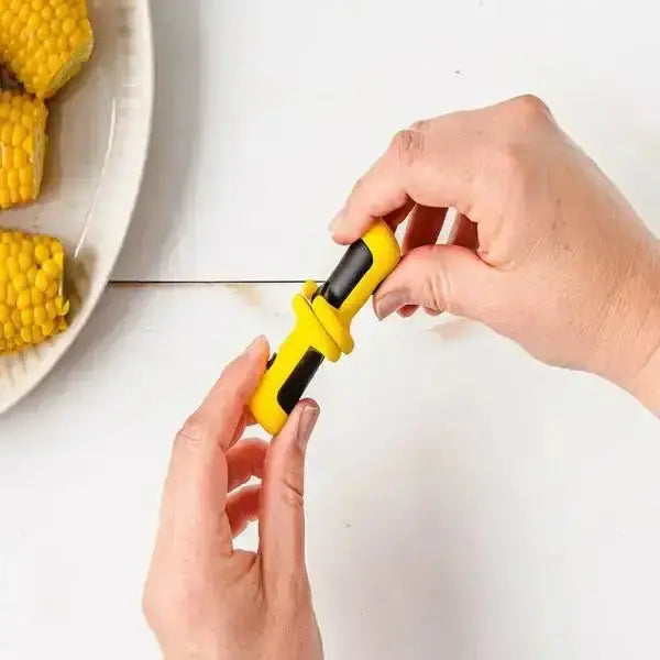 Hands holding Dreamfarm Anchor Corn Lockorns kitchen tool near a plate of corn, Cookin Stores
