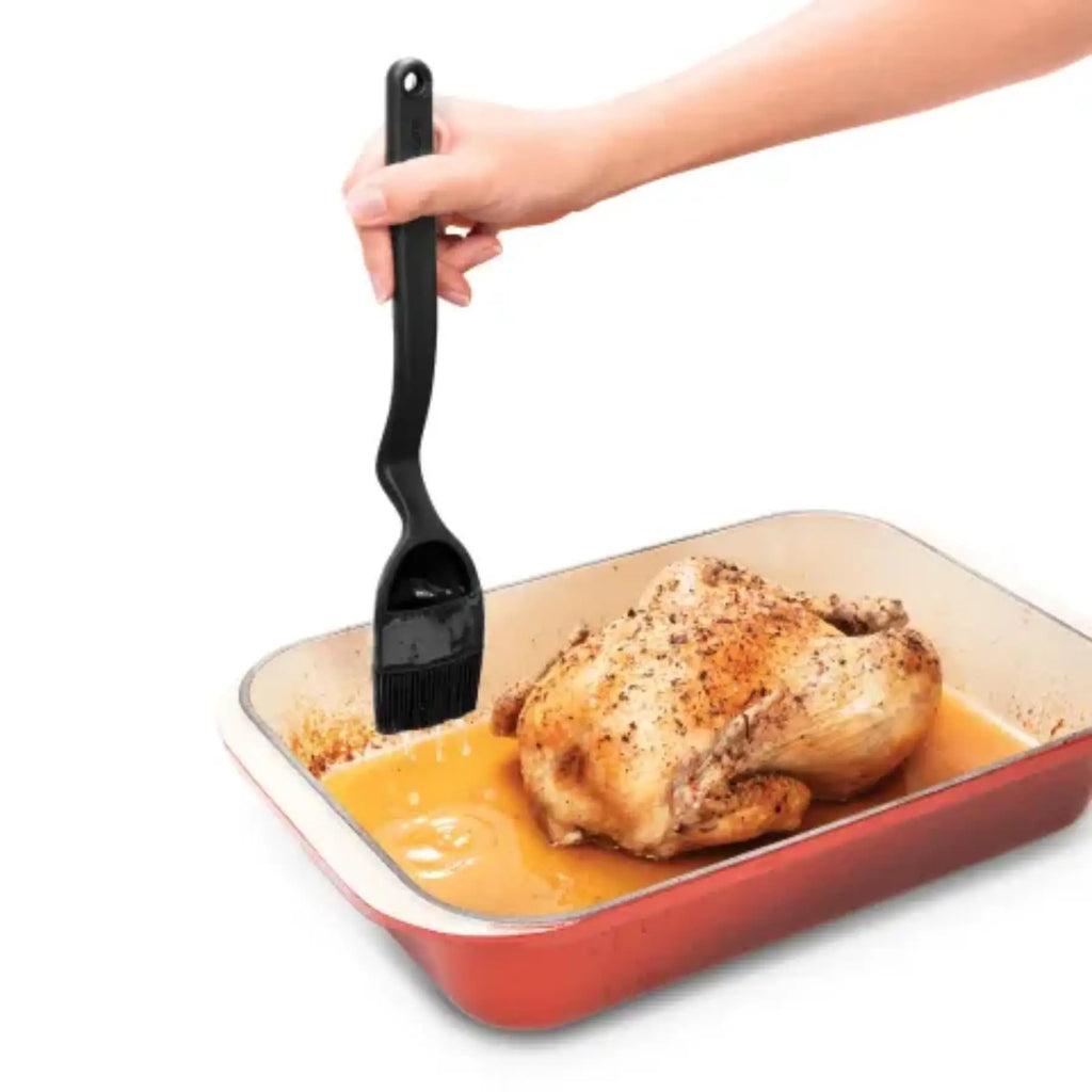 Black dreamfarm bbq brizzle silicone basting brush with long handle applying sauce to roasted chicken in red dish