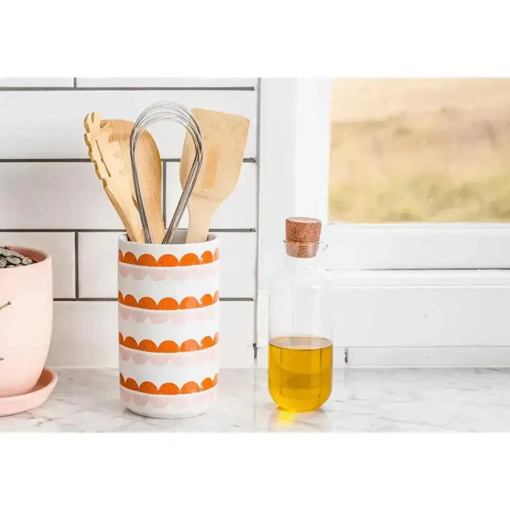Kitchen utensils in a patterned holder next to a glass of yellow liquid on a marble countertop.