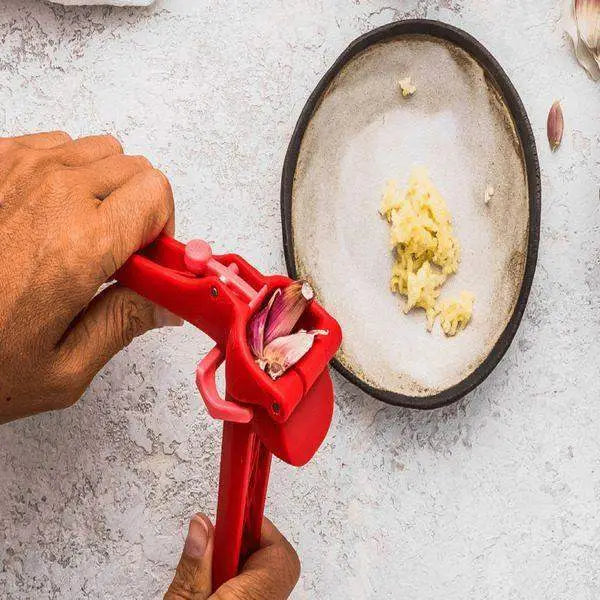 Hand using red garlic press over plate with minced garlic, kitchenware at Cookin Stores