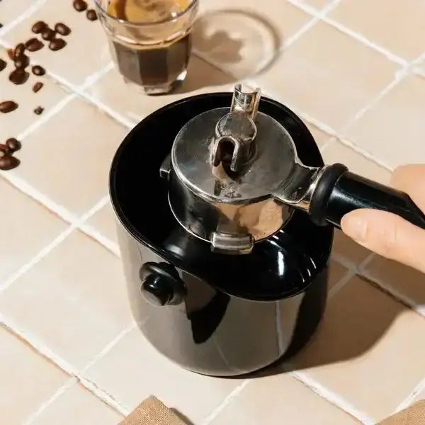 Black coffee grinder with a silver top on a tiled surface, surrounded by coffee beans and a glass of coffee.