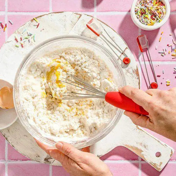 Mixing cake batter in a bowl with a red Dreamfarm Mini Flisk whisk, kitchen tools on board