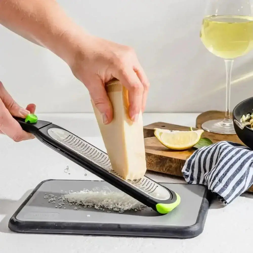 Person using a manual zester on a block of cheese with a glass of white wine in the background.