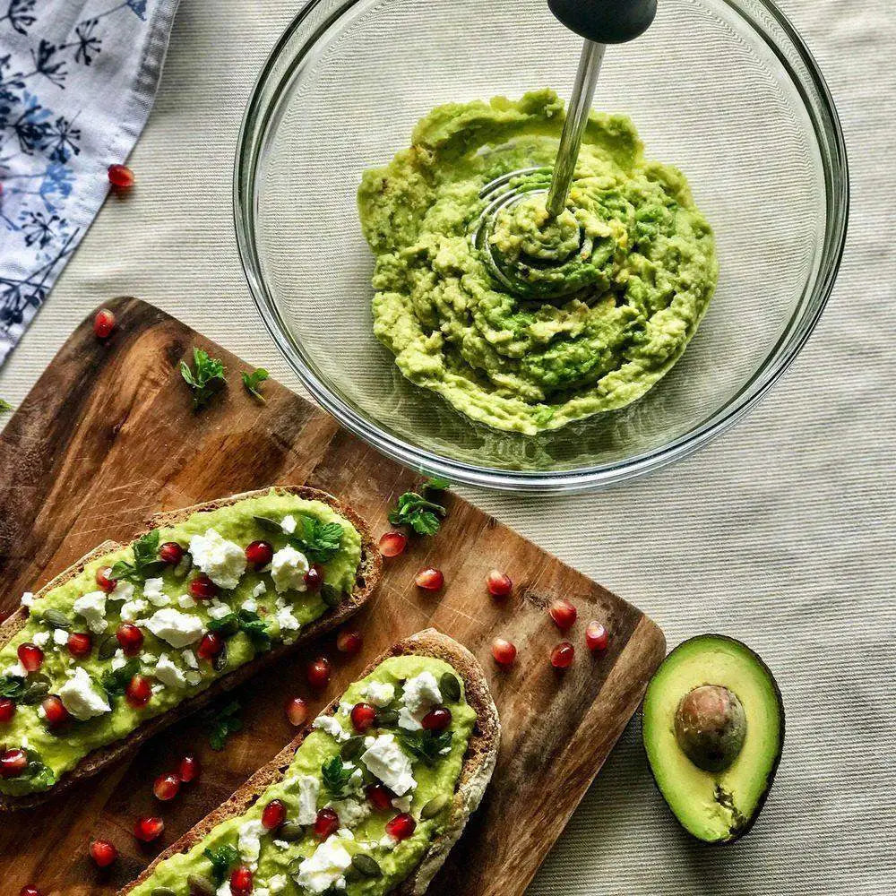 Dreamfarm Smood masher in charcoal black mashing avocado in glass bowl, avocado toast with feta and pomegranate on wood board, kitchenware from Cookin Stores