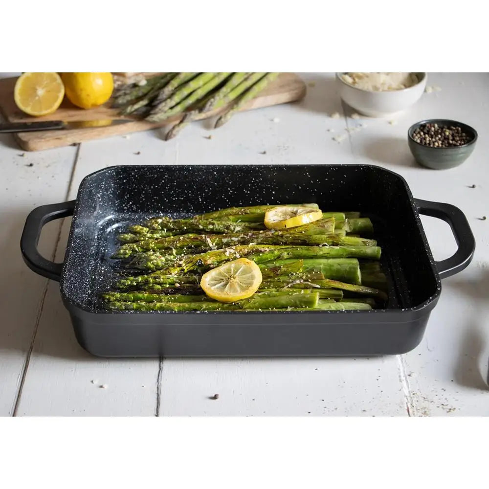 Roasted asparagus with lemon slices in a black speckled baking dish on a white surface.