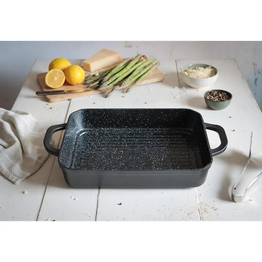 Black speckled rectangular baking dish on a white wooden surface with lemons, asparagus, and other ingredients.