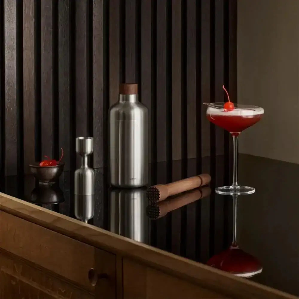 Cocktail with a cherry on a bar counter next to a shaker and other bar tools.