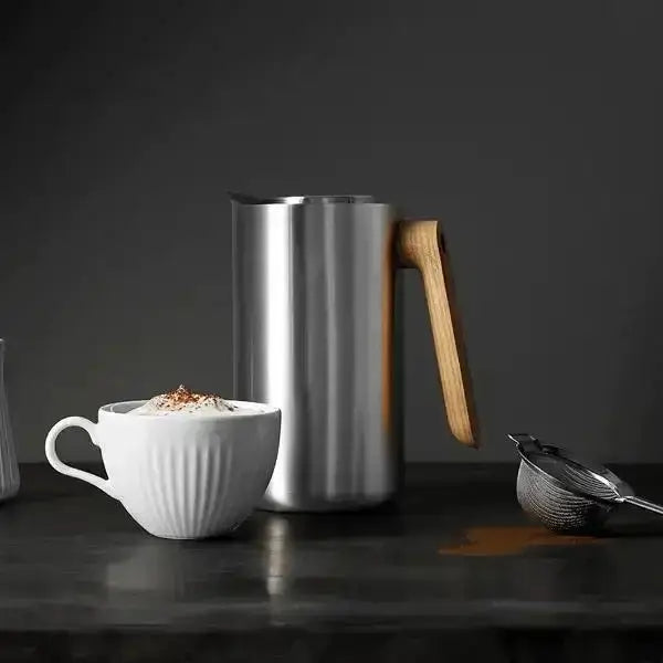 Stainless steel coffee jug with wooden handle, cappuccino cup, and strainer on dark surface at Cookin Stores.