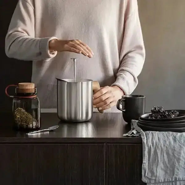 Person preparing a drink using a stainless steel pitcher on a wooden table.