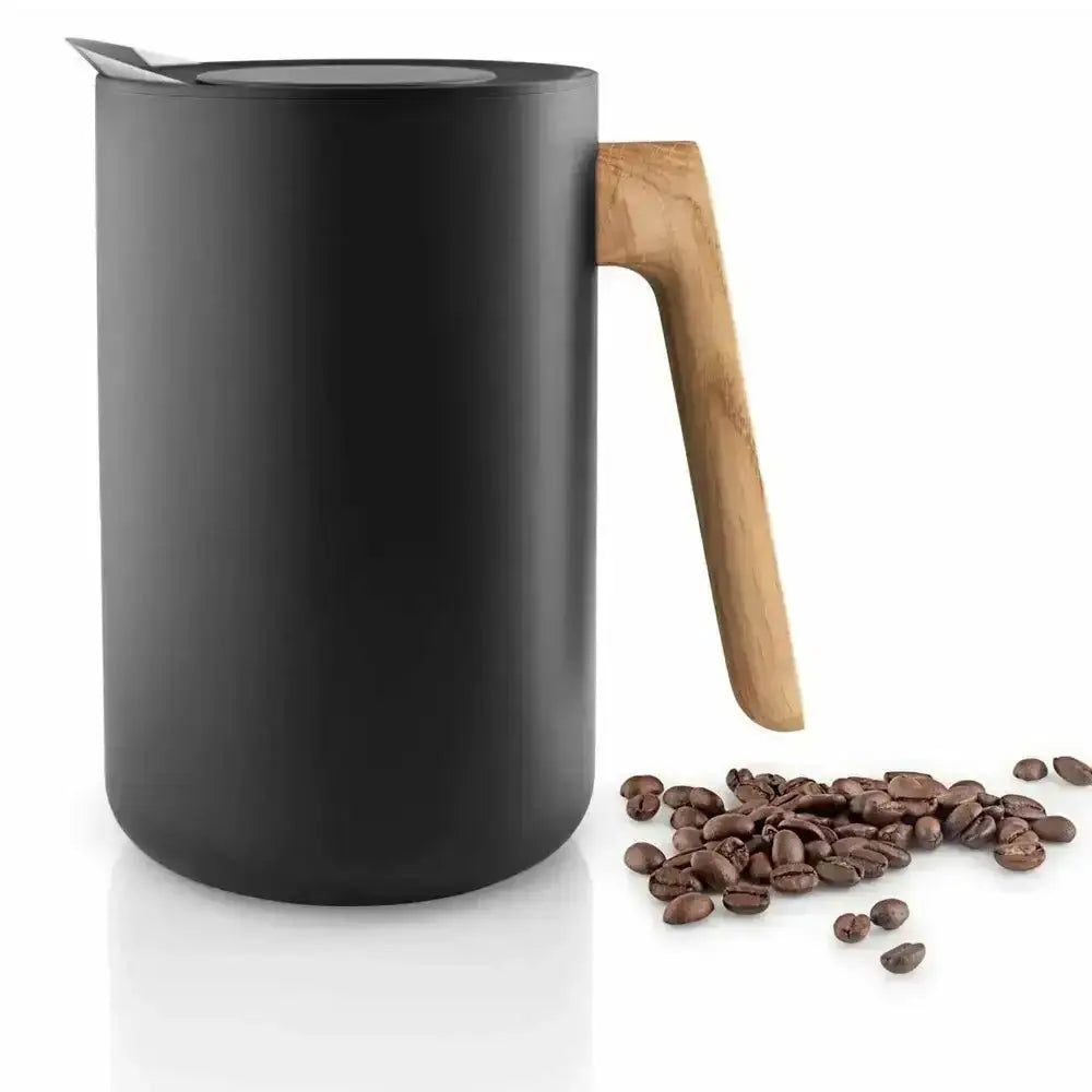 Black milk frothing pitcher with wooden handle next to coffee beans on a white background
