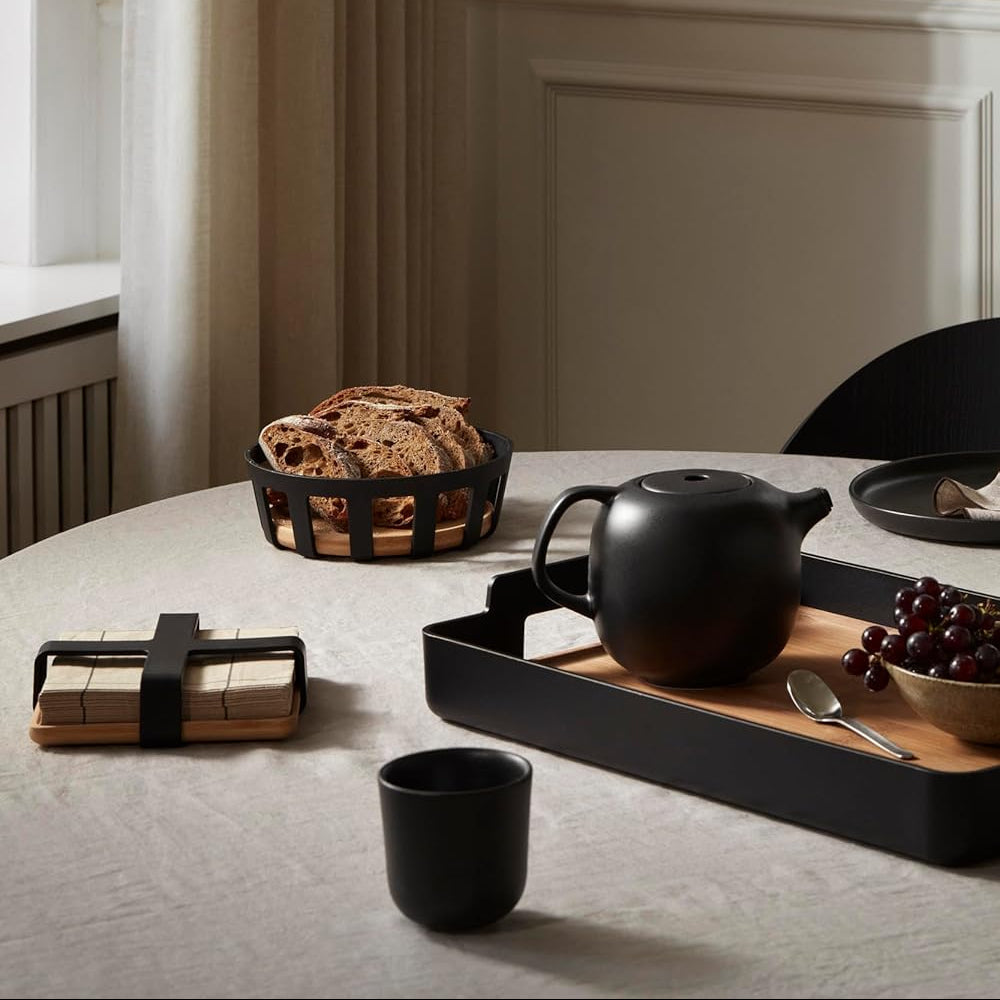 Tea set with a teapot, cups, and bread on a table in a room with curtains and a fireplace.