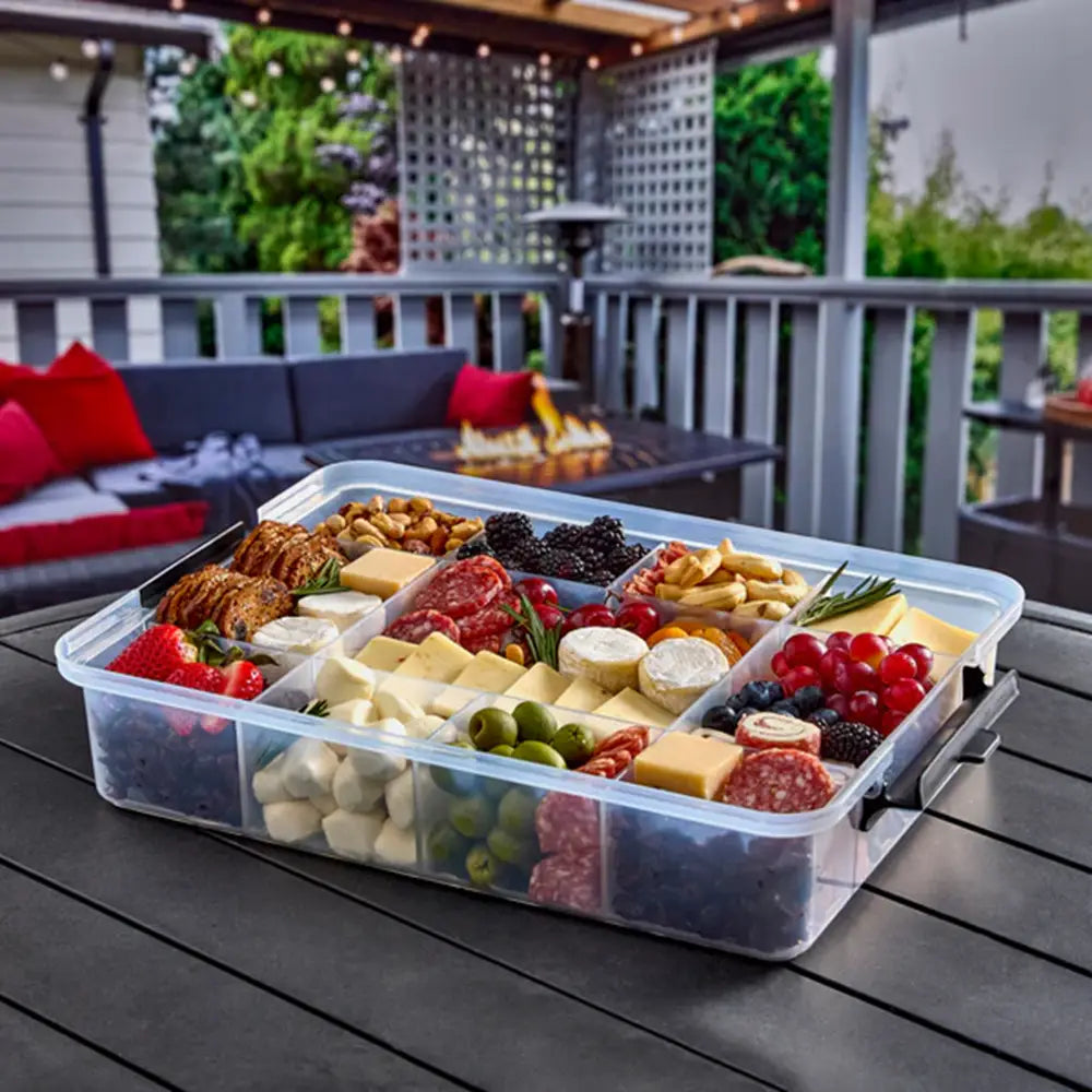Assorted food platter in a clear container on a patio table with outdoor furniture in the background.