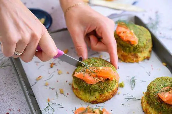 Person slicing savory scone topped with smoked salmon and herbs, on baking tray, kitchenware visible