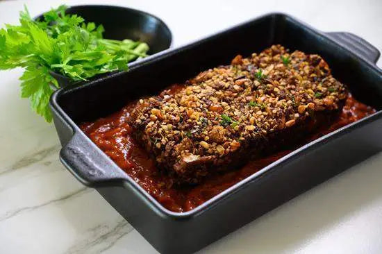 Nut-crusted meatloaf in a black baking dish with tomato sauce, fresh celery nearby, premium kitchenware