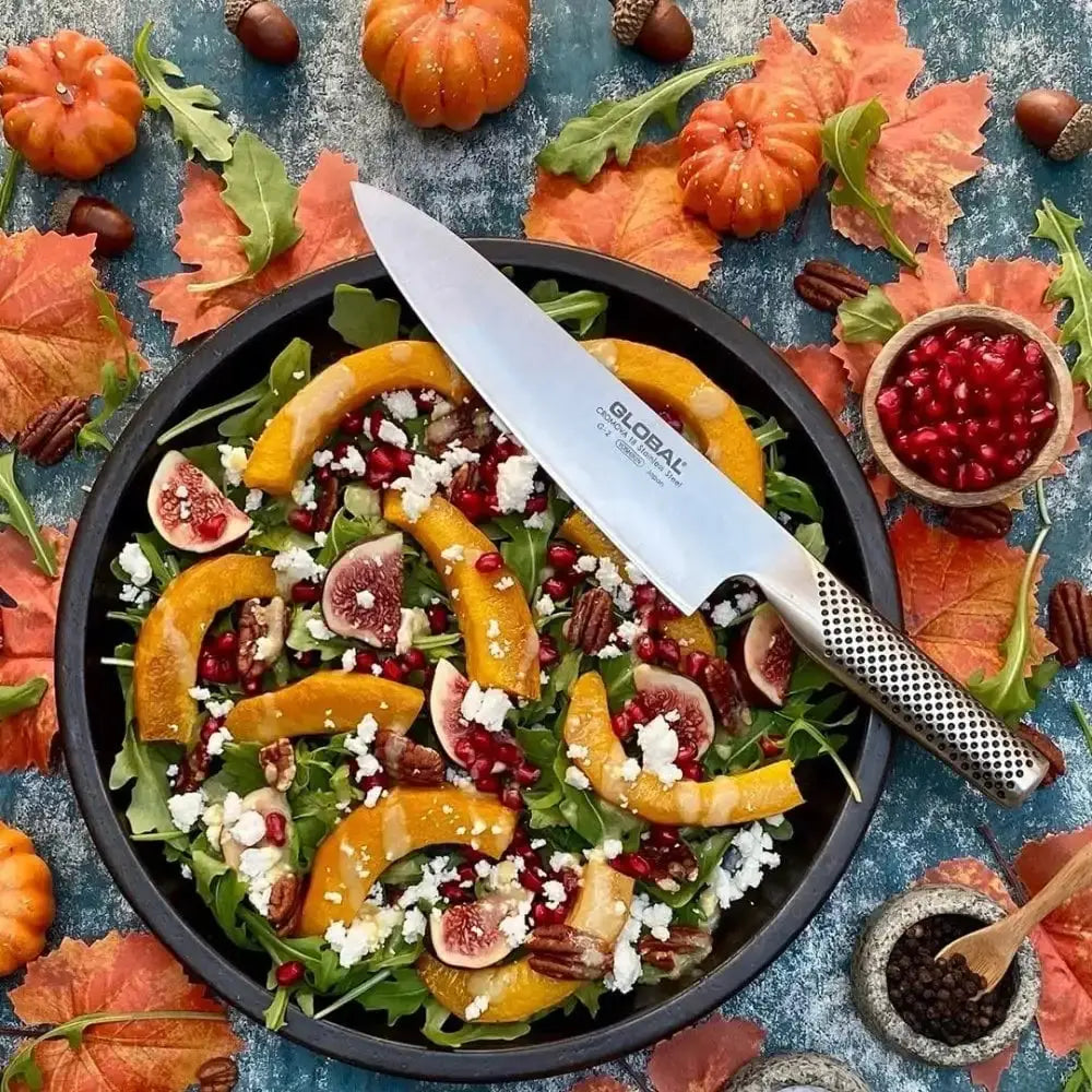 Salad with pumpkins, figs, and pomegranate seeds on a blue surface with autumn leaves and a Global knife.