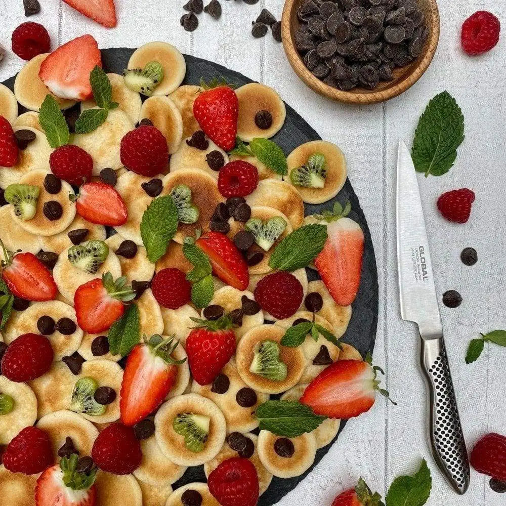 Fruity dessert pizza with strawberries, kiwis, and chocolate chips on a white surface with a knife.