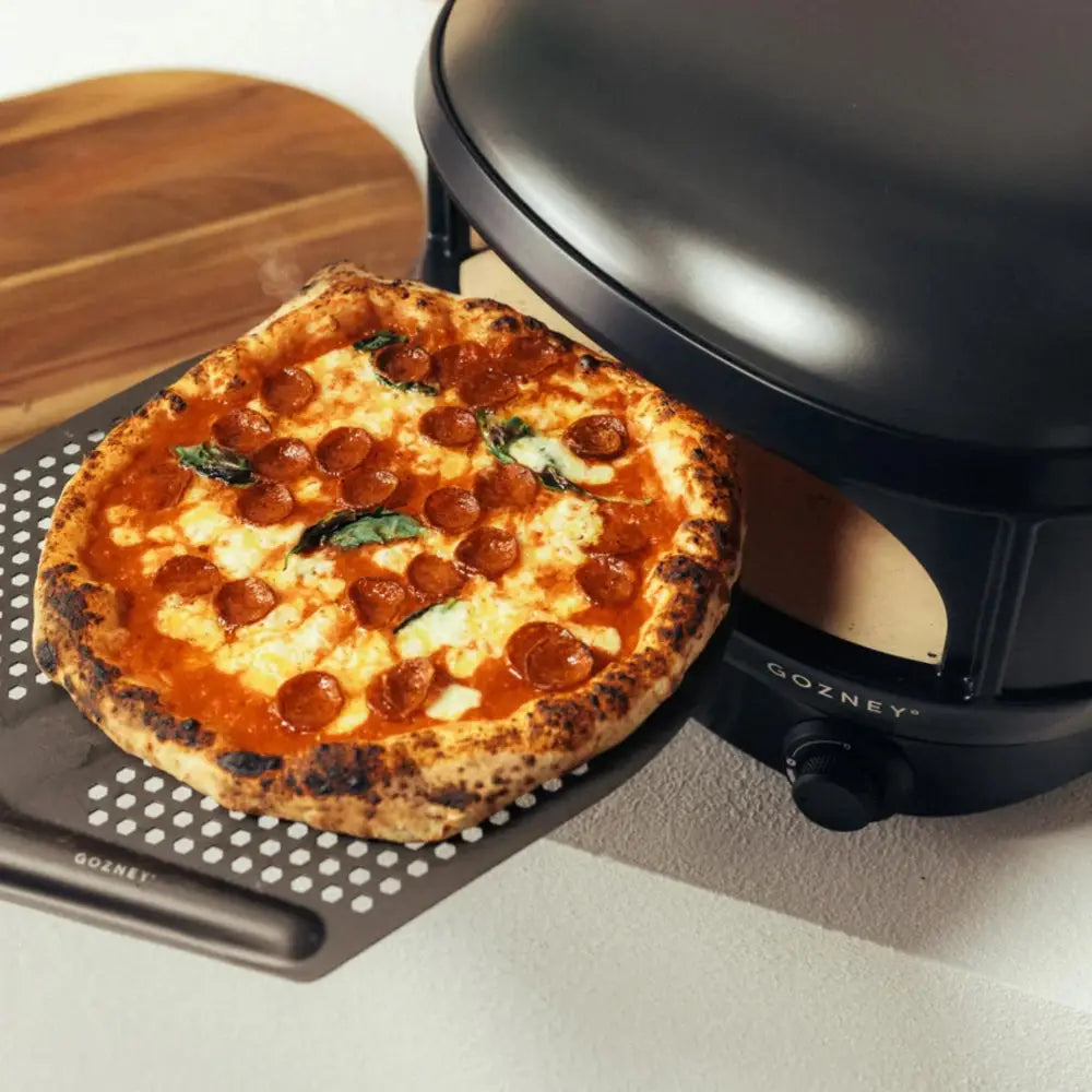 Pepperoni pizza on a Gozney pizza stone with a pizza oven in the background.