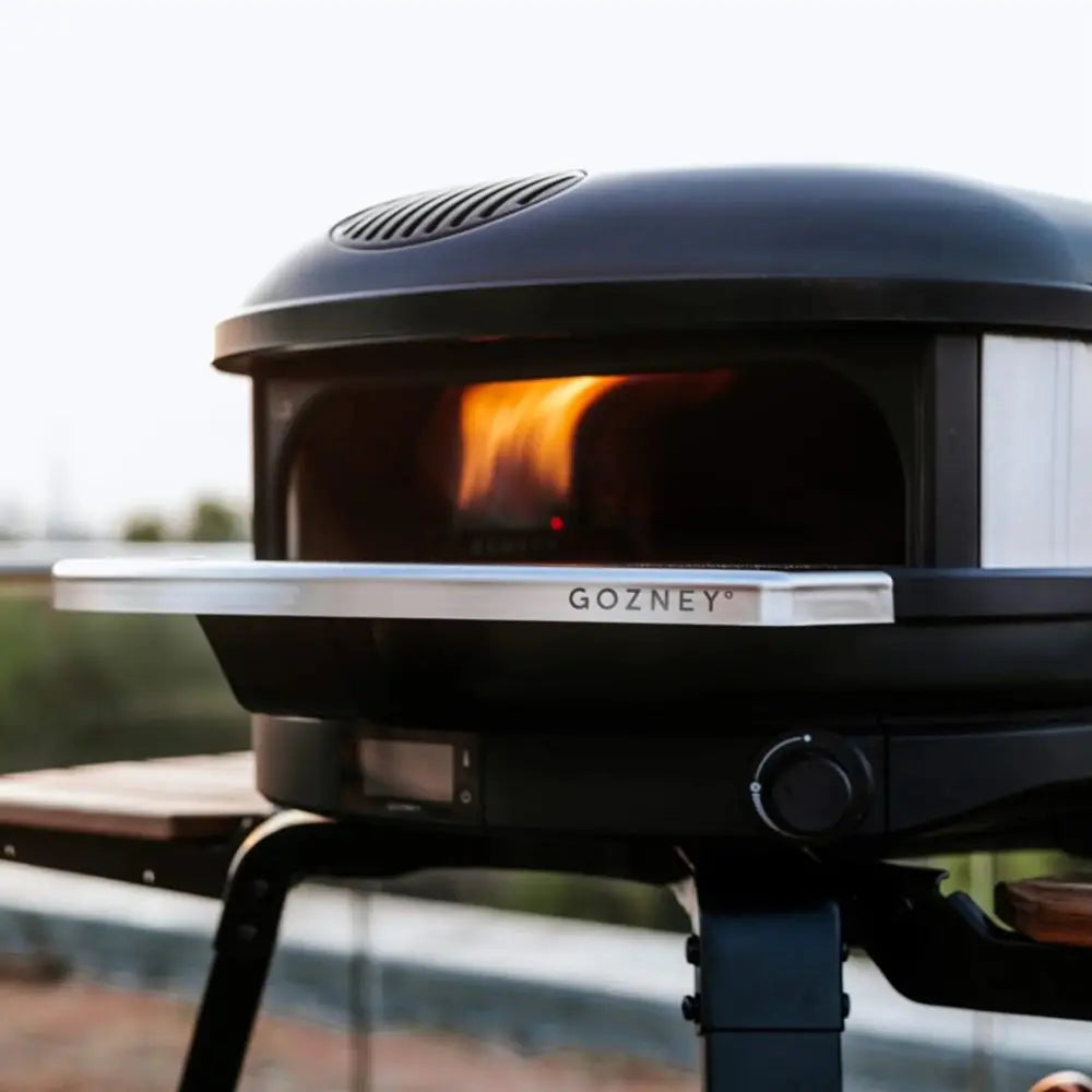 Black Gozney pizza oven with flames inside, outdoors.