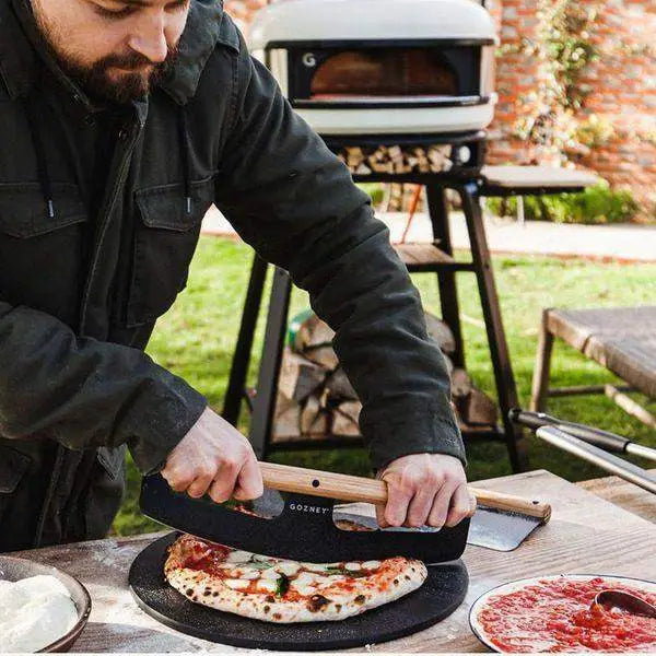 Man slicing pizza on black Gozney pizza server outdoors near wood-fired oven, Cookin Stores kitchenware