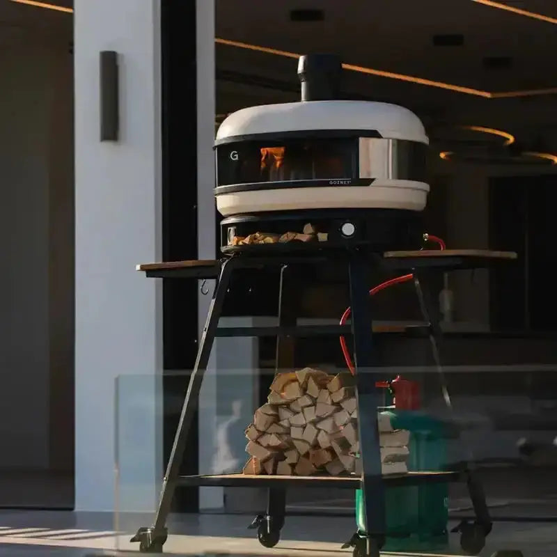 Gozney Dome Bone dual fuel pizza oven on wheeled stand with wood and gas tank outdoors