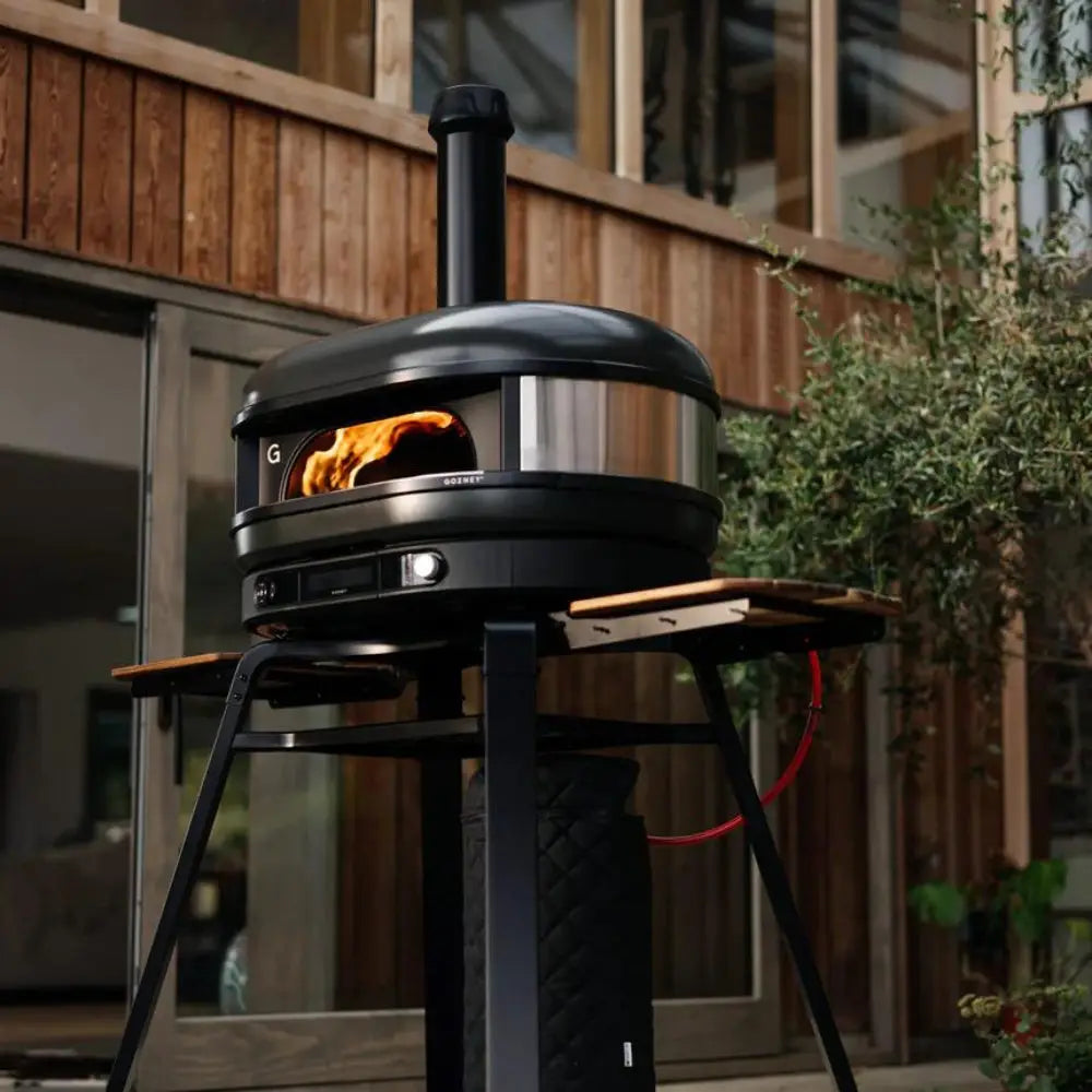 Portable pizza oven on a stand with flames inside, set against a wooden deck background.
