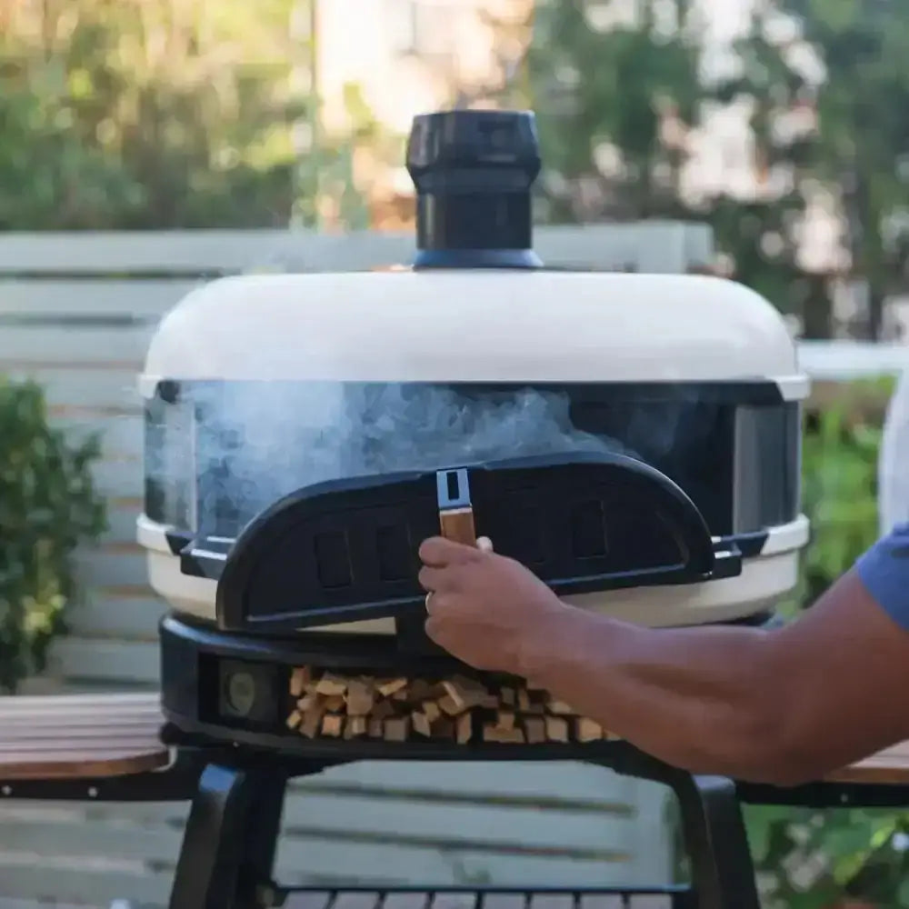 Outdoor Gozney Dome pizza oven with rope-sealed door, wood storage, and visible smoke