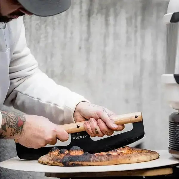 Person using Gozney pizza cutter rocker with wooden handle to slice fresh pizza, Cookin Stores kitchenware