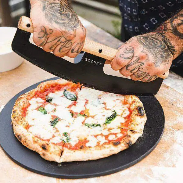 Person slicing Margherita pizza with Gozney pizza cutter rocker on wood table, Cookin Stores kitchenware.