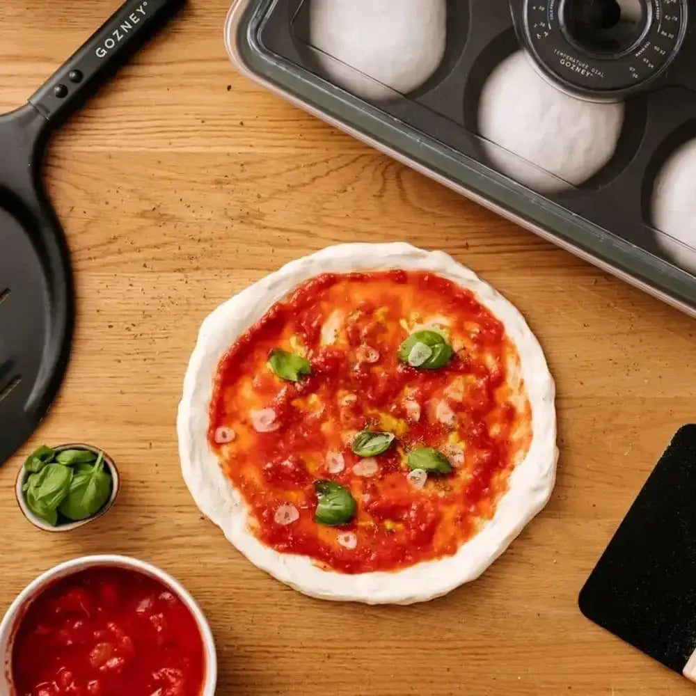Small pizza with tomato sauce and basil leaves on a wooden surface, with a muffin tin and ingredients nearby.