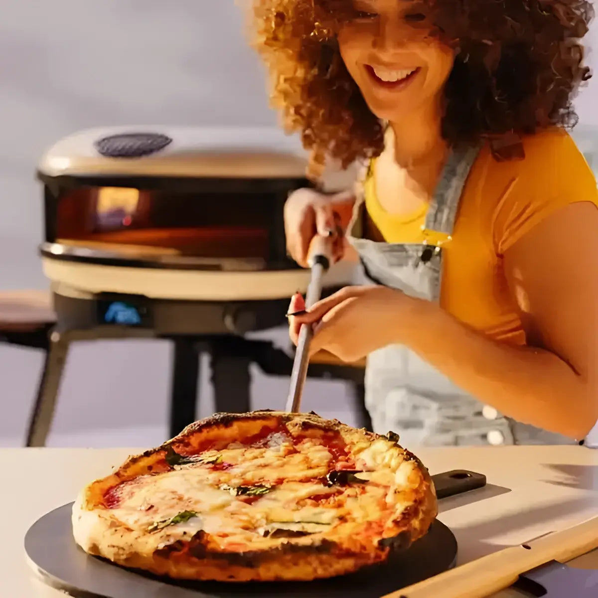Smiling woman using a Gozney pizza peel with a fresh pizza, outdoor pizza oven in background
