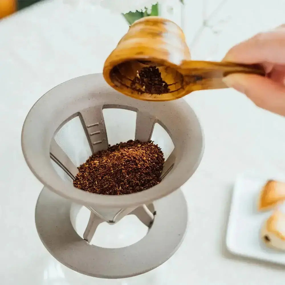 Person using a wooden scoop to add ground coffee into a metal filter.