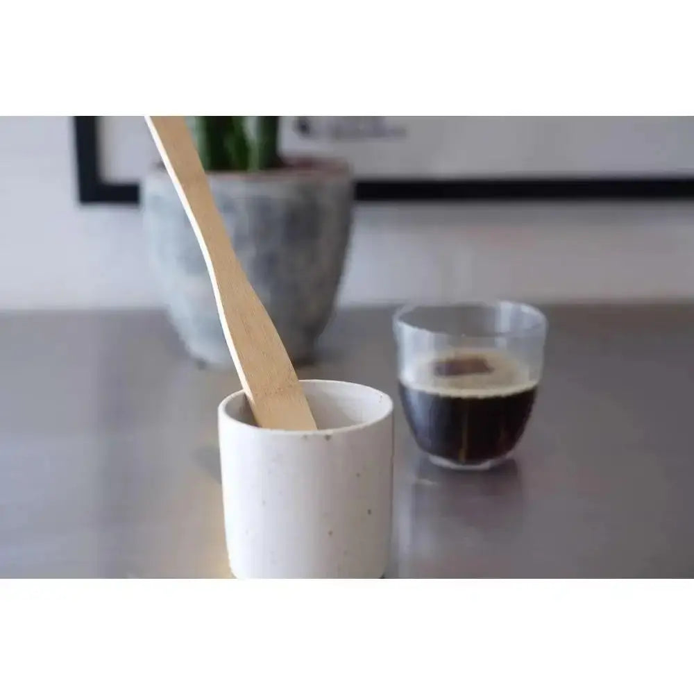 White ceramic cup with a wooden spoon on a kitchen counter, with a glass of coffee in the background.