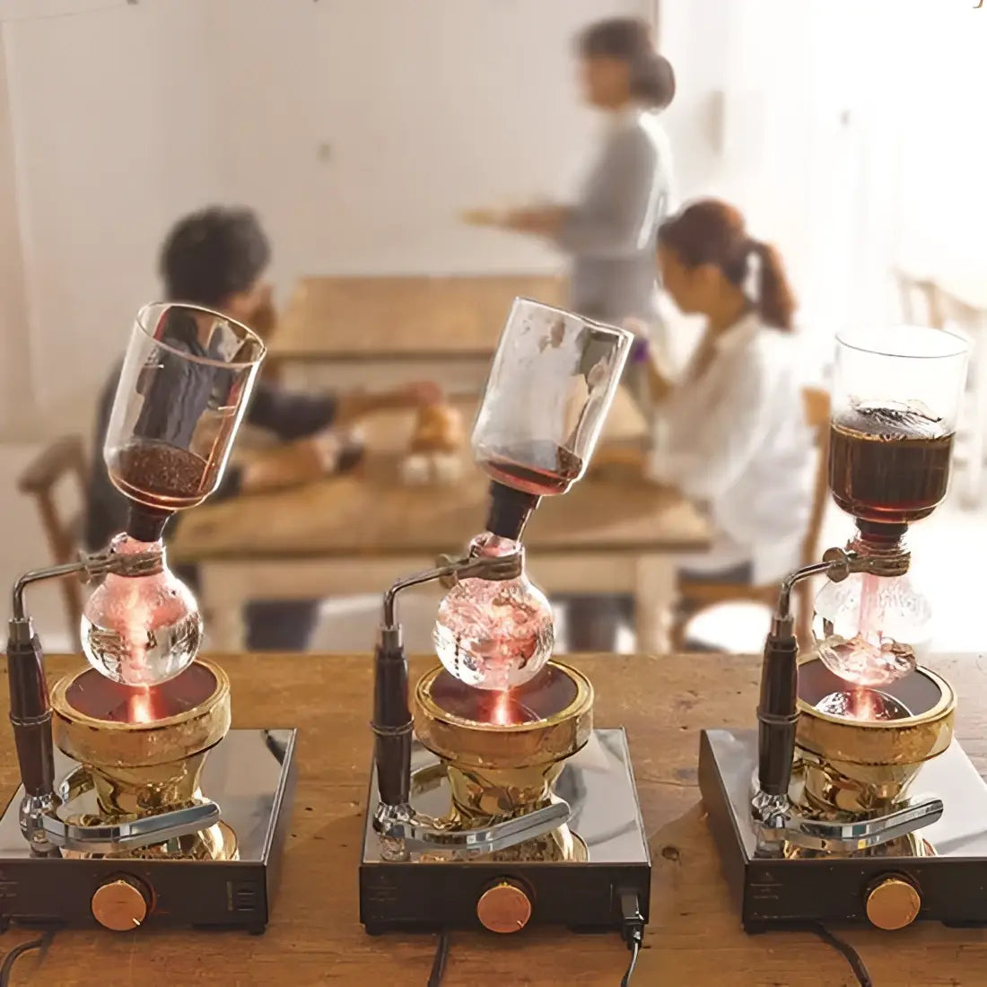 Three siphon coffee apparatuses on a wooden table with people in the background.