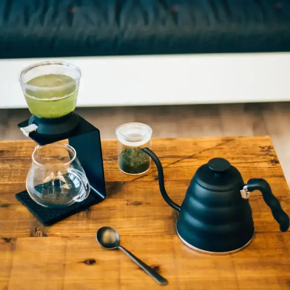 Coffee brewing setup with a kettle, glass carafe, and green liquid on a wooden surface.