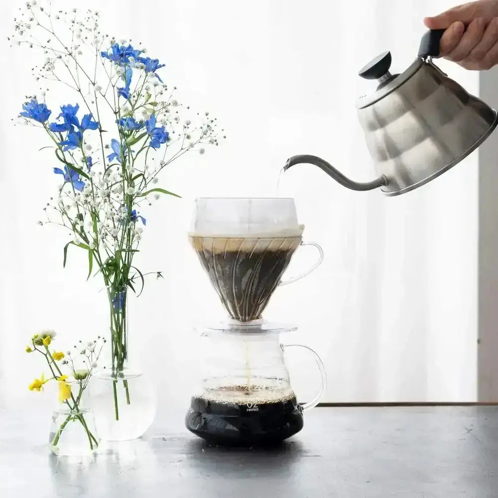 Person pouring coffee from a kettle into a pour-over coffee maker with flowers in the background.