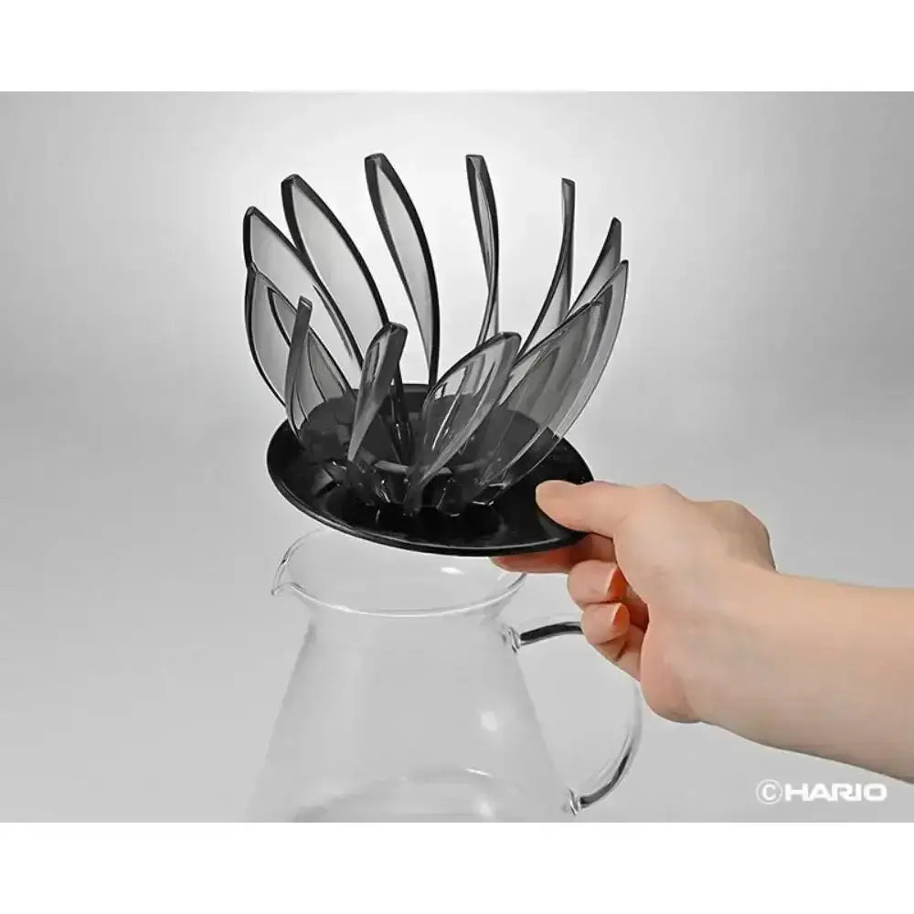 Hand holding a black Hario coffee filter basket over a clear glass coffee pot on a light gray background.