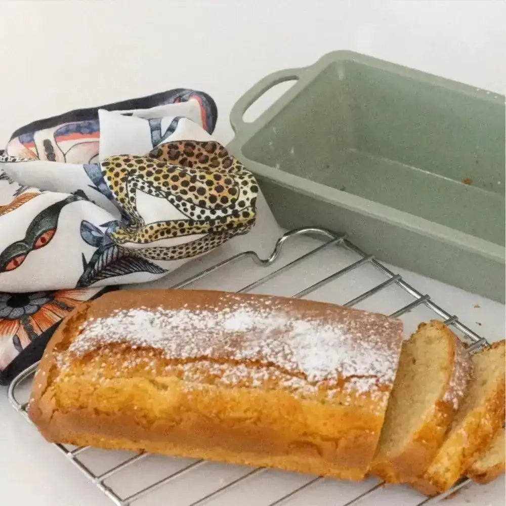 Loaf of bread with powdered sugar on a cooling rack with baking pans and cloths in the background.
