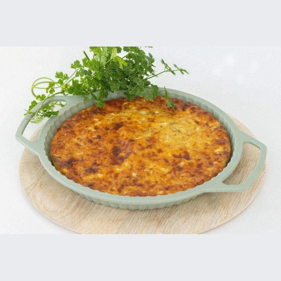 Silicone round baking pan with handles holding baked dish, topped with fresh herbs on a wooden board