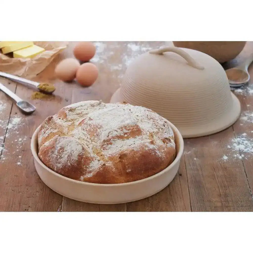 Loaf of bread in a ceramic dish on a wooden surface with flour and eggs.