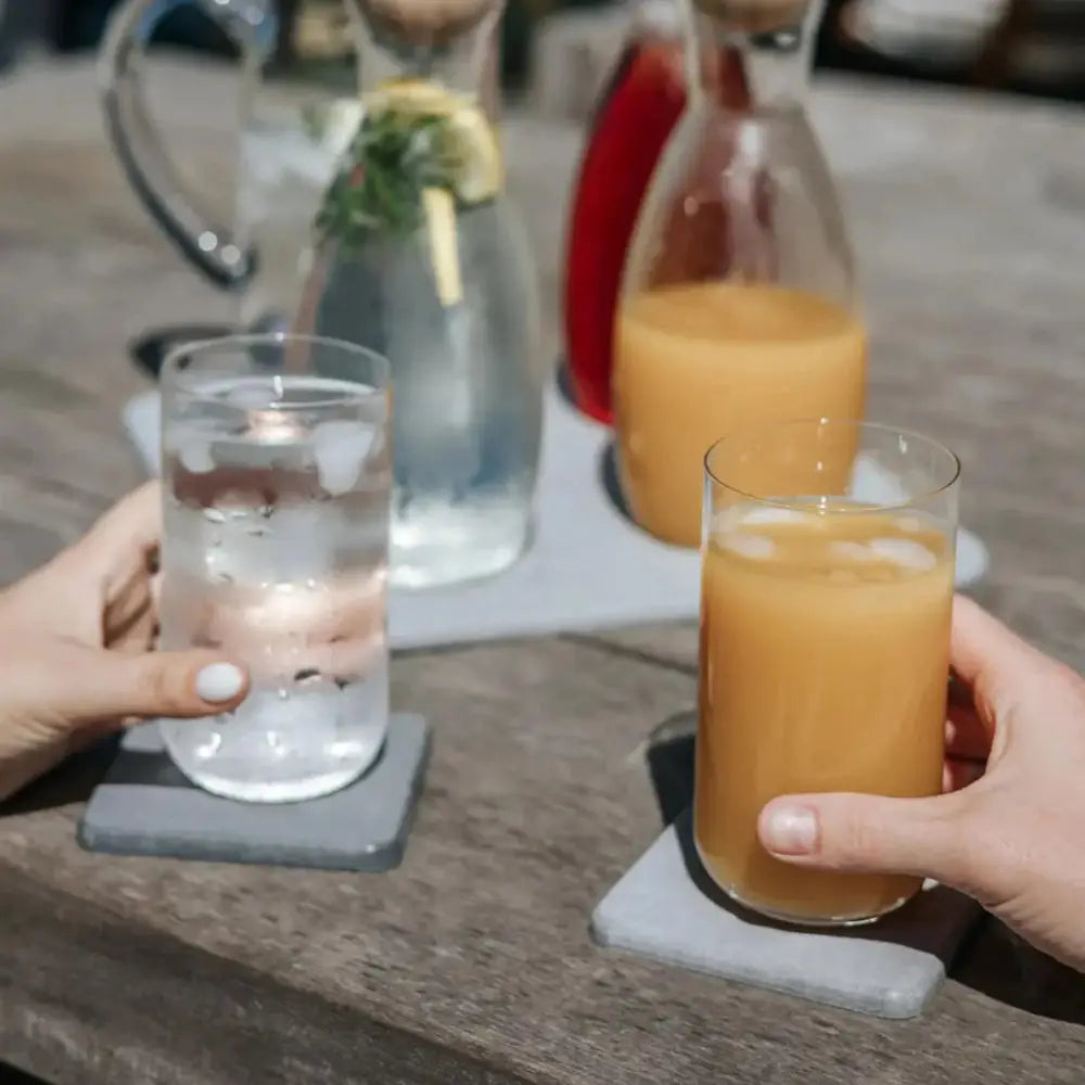 Two hands holding glasses of water and orange juice on a wooden table.