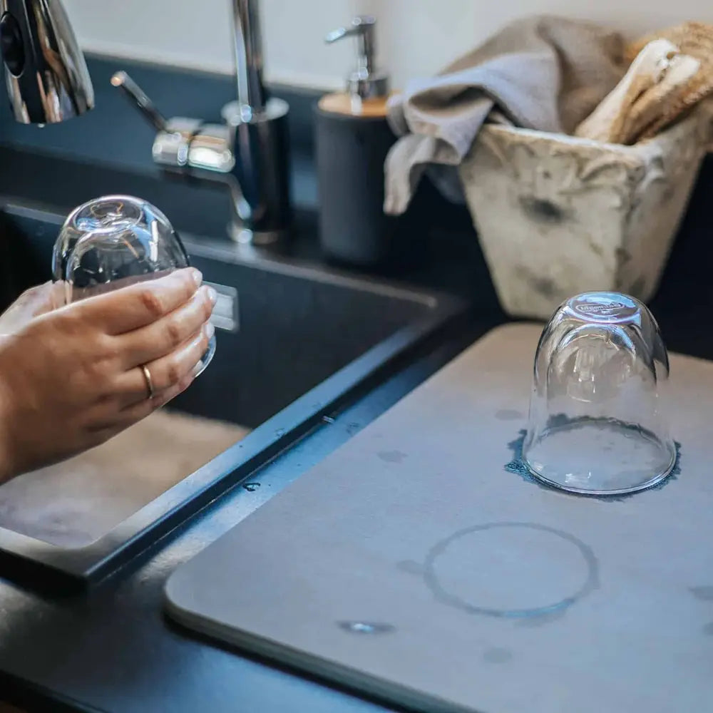 Person washing a glass in a kitchen sink with a drying rack and utensil basket in the background.