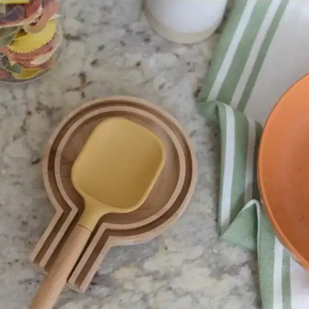 Yellow spatula on a wooden holder with an orange plate and striped towel in the background.